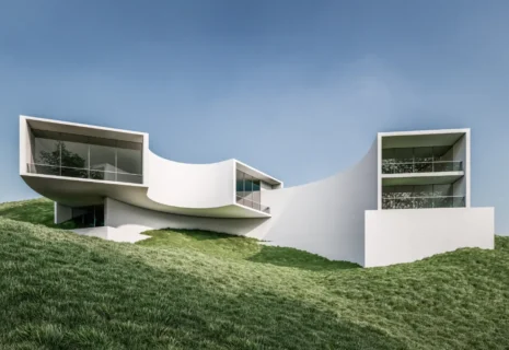 White modern hillside house with curved walls and large glass windows under a clear blue sky.