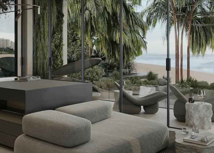 Modern living area with a gray daybed and built-in platform, glass walls, and a beachside view with palm trees outside.