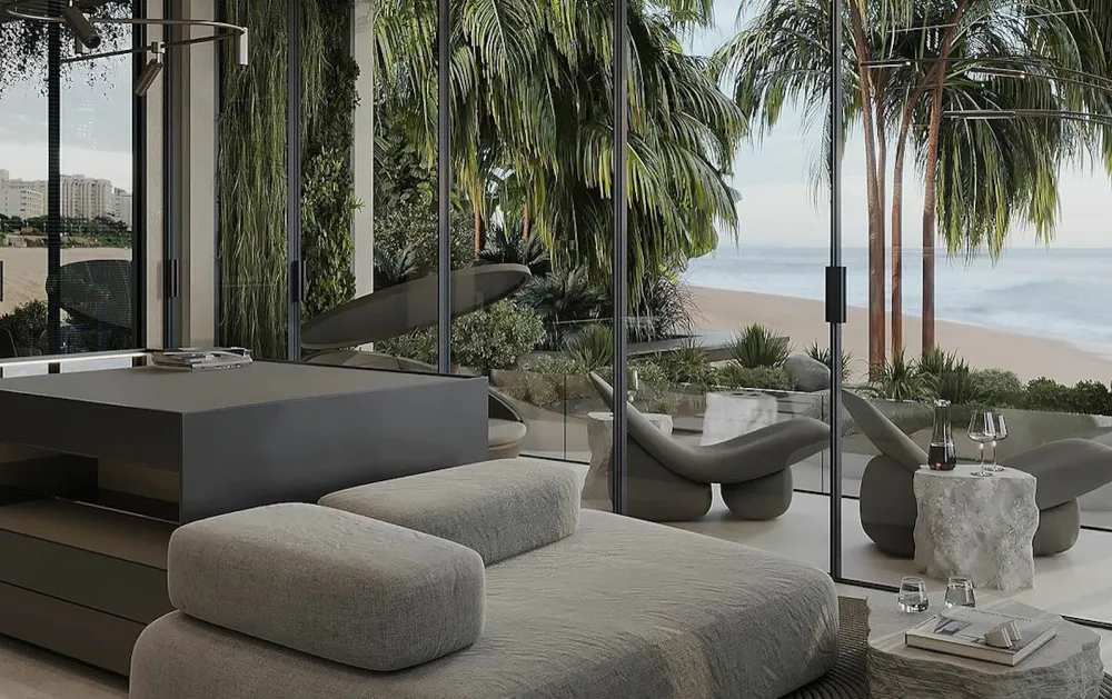 Modern living area with a gray daybed and built-in platform, glass walls, and a beachside view with palm trees outside.
