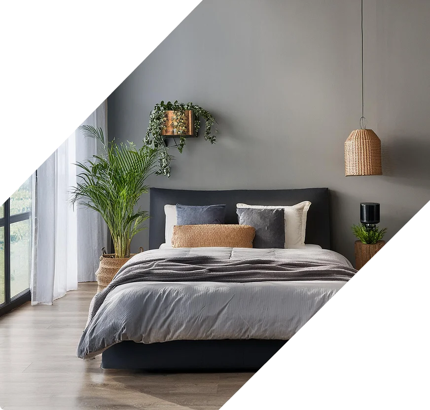 Modern bedroom with grey bedding, dark headboard, and lush potted plants by a large window with sheer curtains.