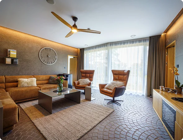 Modern living room with brown leather sofa, two armchairs, glass coffee table, large sheer-curtained windows, and a wall clock on a textured wall, illuminated by warm light.