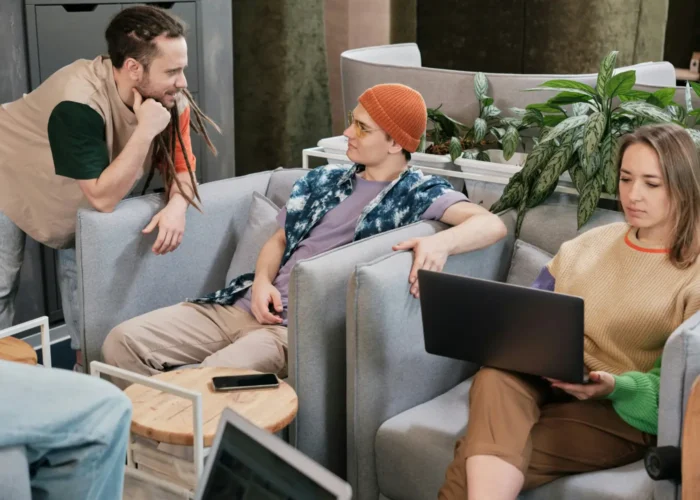 Three friends in a contemporary living room: one man with dreadlocks leaning on a sofa, another man in an orange beanie lounge chatting, while a woman on the right types on a laptop with plants in the background.