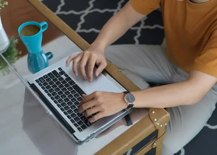 Person typing on a silver laptop at a glass coffee table, with a blue mug nearby.
