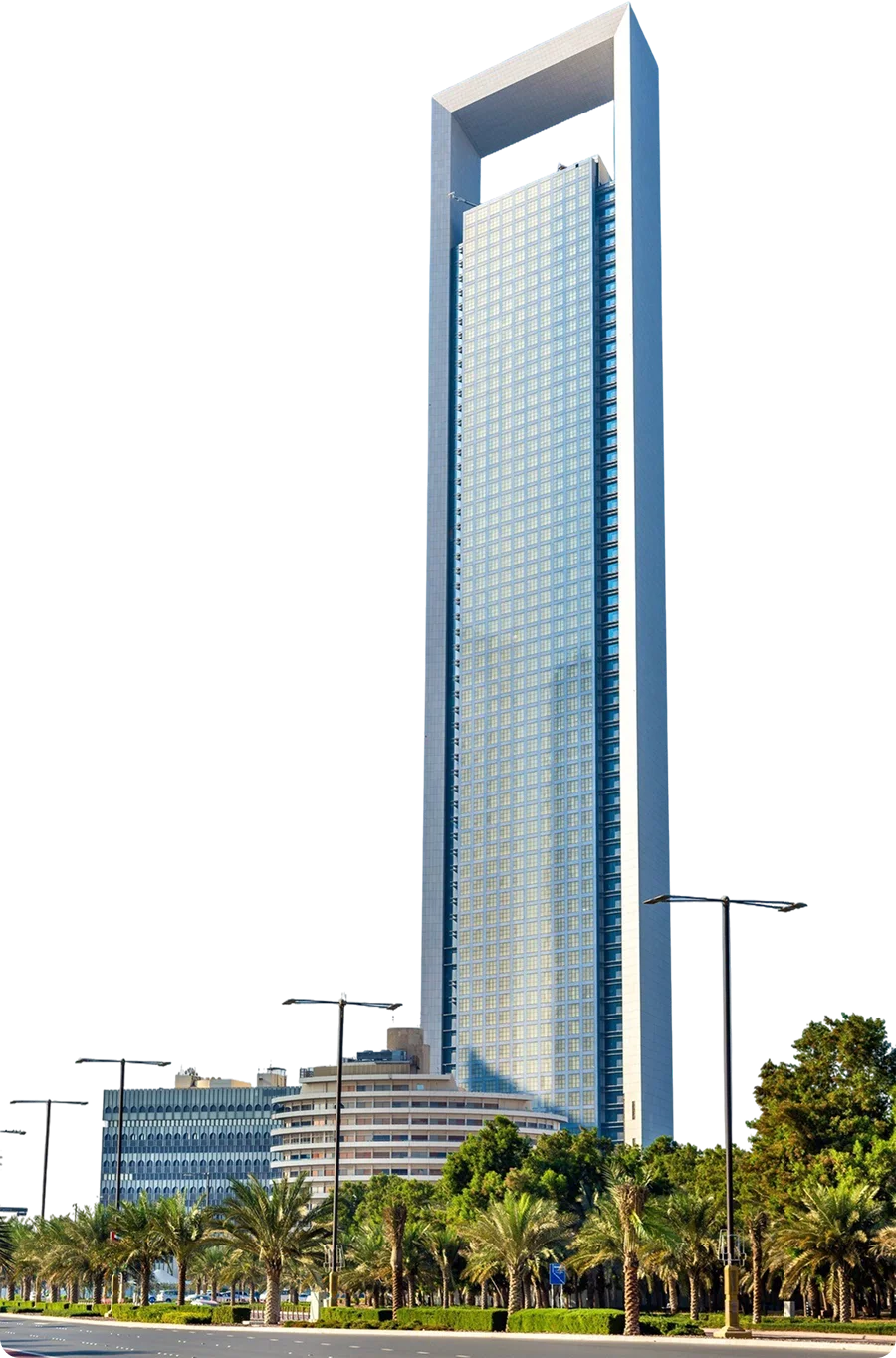img-about-h3 - beachfrontcondopattaya Tall modern glass skyscraper with a square arch at the top, standing along a palm-lined street with streetlights and smaller buildings nearby.
