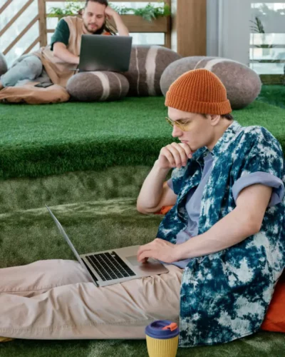Young man in an orange beanie uses a laptop on artificial grass with a takeaway cup beside him; a coworker lounges on bean bags in the background.