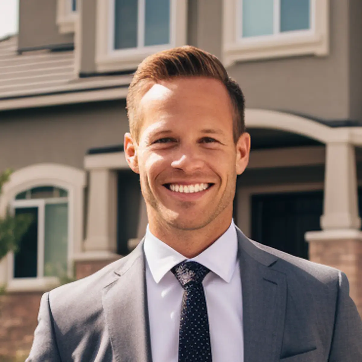 Smiling man in a gray suit and polka-dot tie outdoors in front of a house.