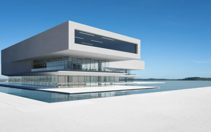 Futuristic white multi-story building with glass walls sits over a reflecting pool by calm water under a clear blue sky.