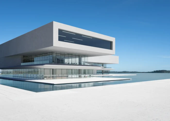 Futuristic white multi-story building with glass walls sits over a reflecting pool by calm water under a clear blue sky.