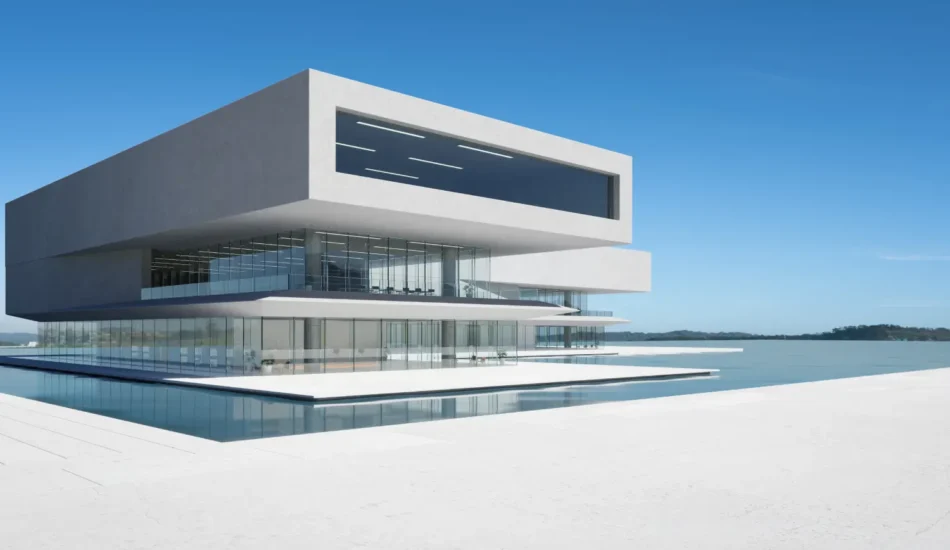Futuristic white multi-story building with glass walls sits over a reflecting pool by calm water under a clear blue sky.