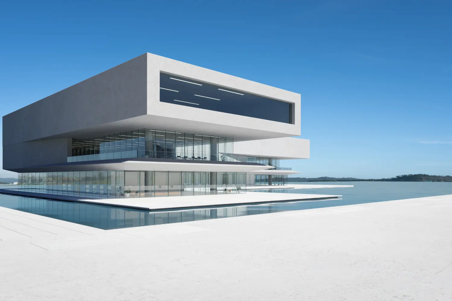 Futuristic white multi-story building with glass walls sits over a reflecting pool by calm water under a clear blue sky.