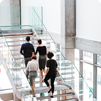 Four people ascend a glass staircase in a bright, modern building with large windows on the sides.