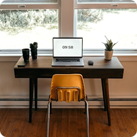 Wooden desk by a window with a laptop, two potted plants, and an orange chair in front of it.